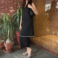 Woman in a black shalwar kameez standing next to potted plants against a brick wall.