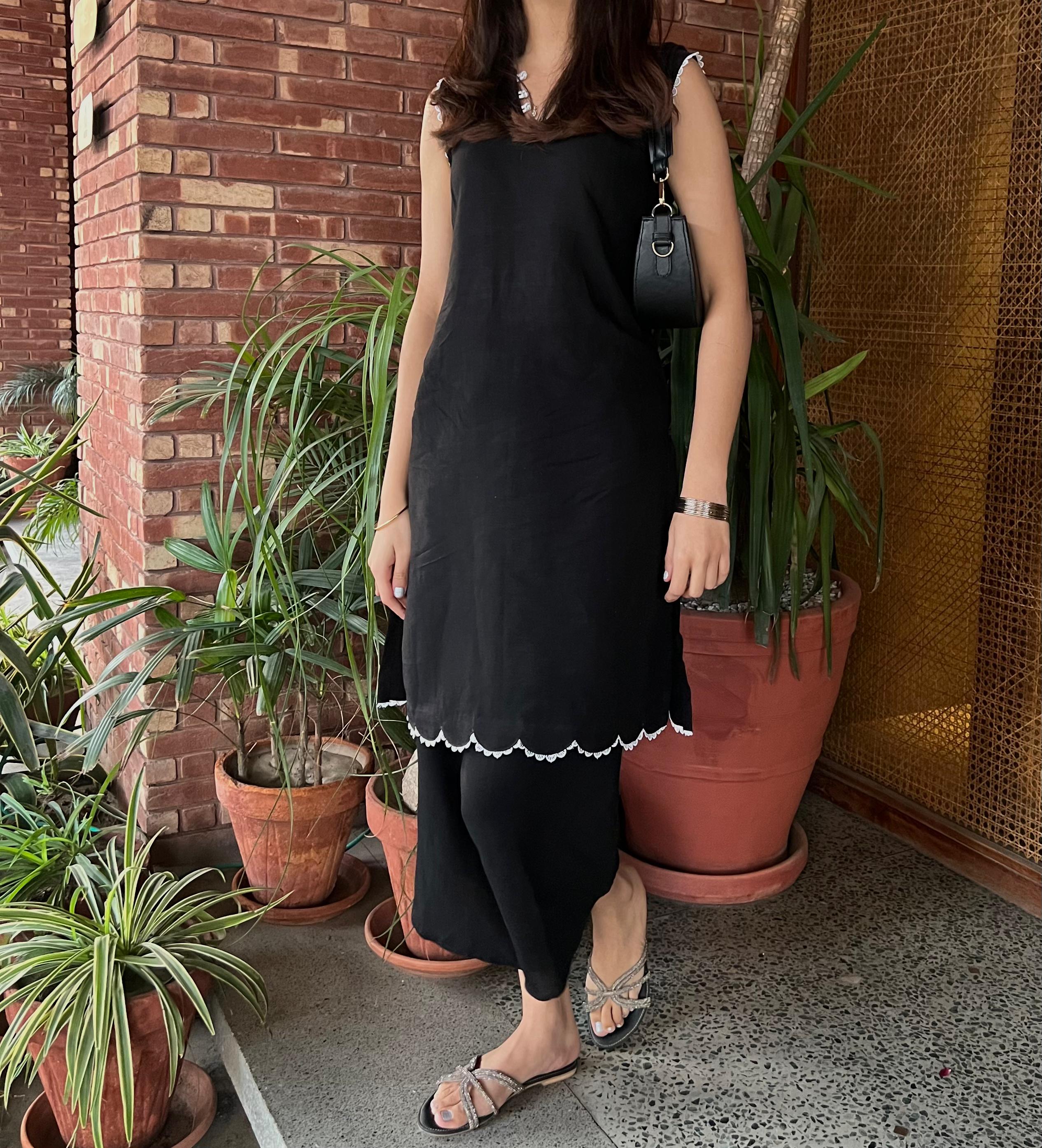 Person wearing a black shalwar kameez standing among potted plants against a brick wall.