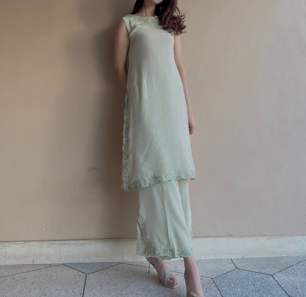 Woman wearing a light green dress with lace details against a plain wall.
