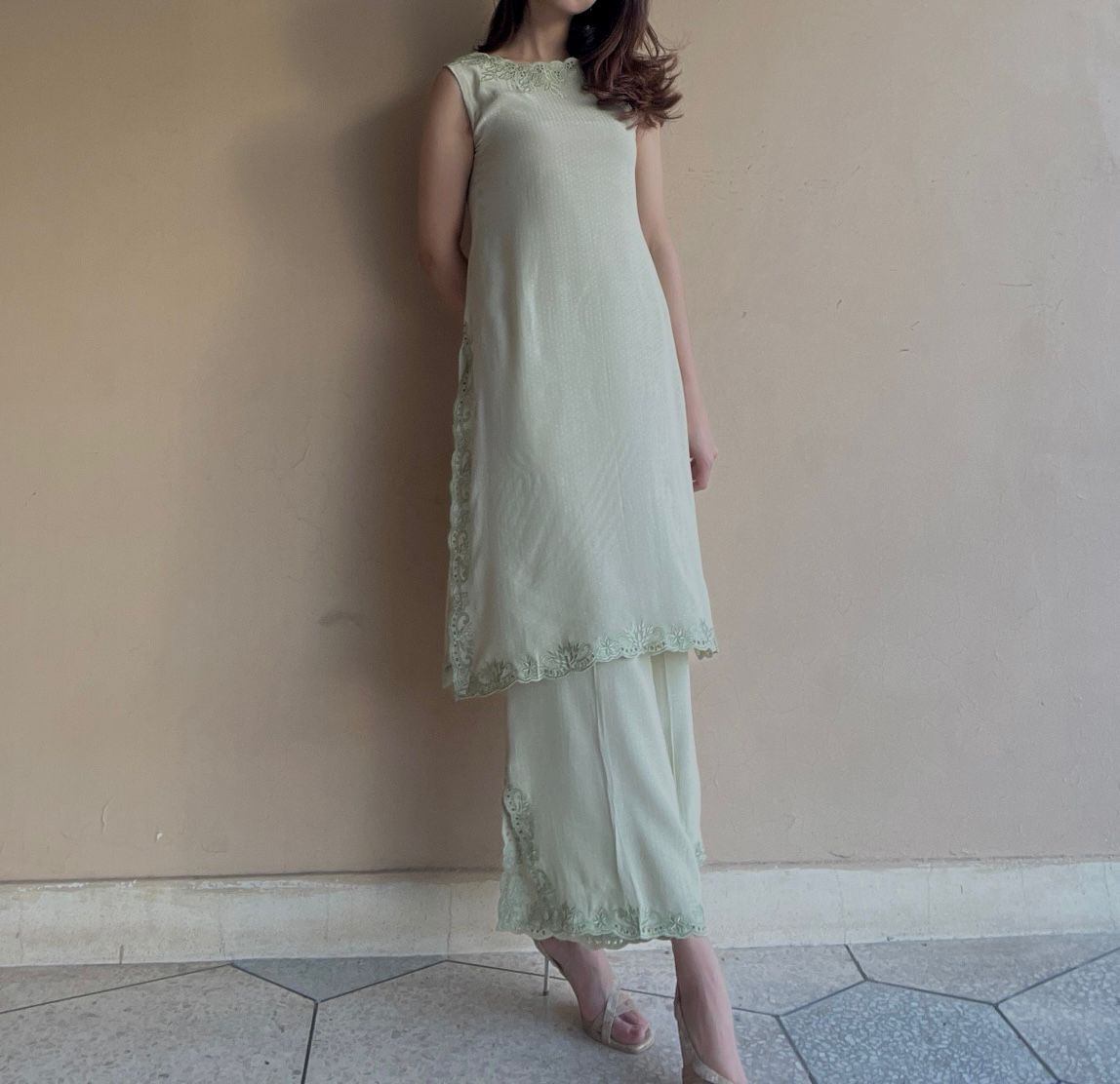 Woman wearing a light green dress with lace details against a plain wall.